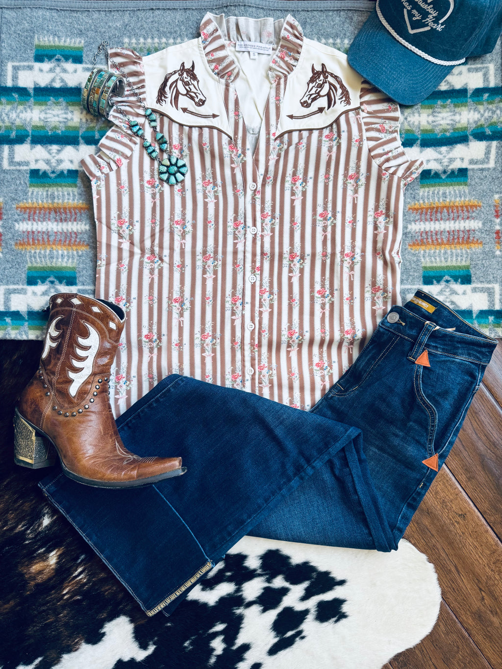A styled flat-lay outfit featuring a sleeveless western-style top with vertical pink and beige stripes, embroidered horse motifs on the shoulders, paired with dark blue jeans, a brown leather cowboy boot, and a teal baseball cap, arranged on a patterned rug and wood floor.