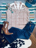 A styled flat-lay outfit featuring a sleeveless western-style top with vertical pink and beige stripes, embroidered horse motifs on the shoulders, paired with dark blue jeans, a brown leather cowboy boot, and a teal baseball cap, arranged on a patterned rug and wood floor.