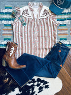 A styled flat-lay outfit featuring a sleeveless western-style top with vertical pink and beige stripes, embroidered horse motifs on the shoulders, paired with dark blue jeans, a brown leather cowboy boot, and a teal baseball cap, arranged on a patterned rug and wood floor.