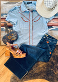 A styled flat-lay outfit featuring a light blue western short-sleeve button-up shirt with orange-brown contrast stitching, dark blue jeans, brown platform sandals, a navy embroidered crossbody bag, and a beige wide-brim hat, arranged on a patterned rug and cowhide rug over a wooden floor.