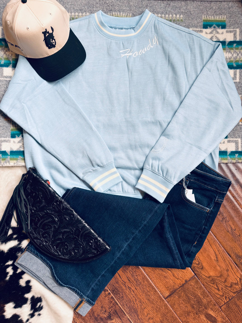 Light blue sweatshirt with Varsity Rodeo branding, dark jeans, black embroidered bag, and beige cap on a wooden floor.