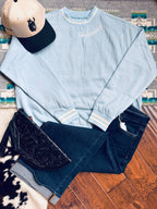 Light blue sweatshirt with Varsity Rodeo branding, dark jeans, black embroidered bag, and beige cap on a wooden floor.