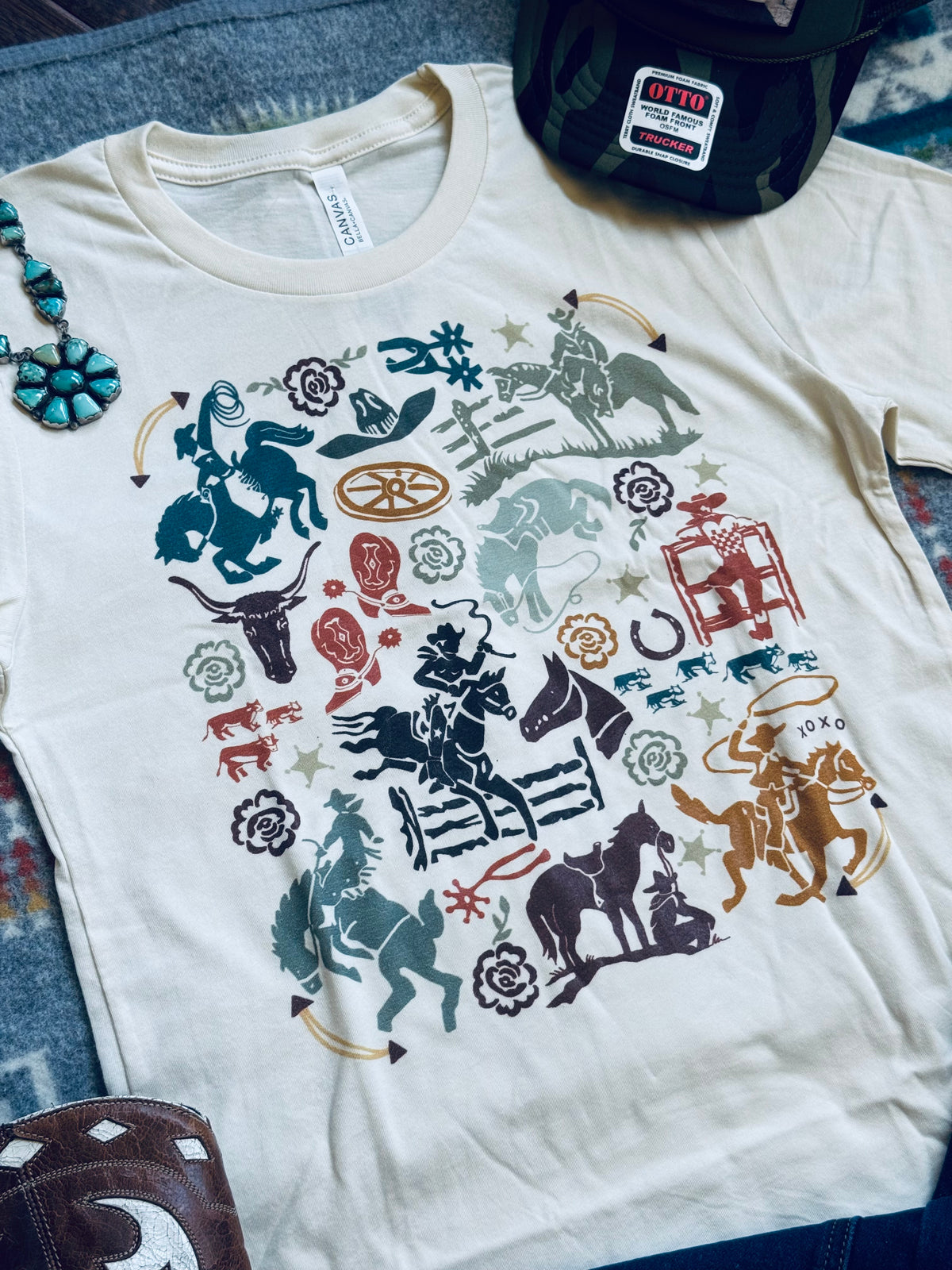 An off-white crewneck t-shirt featuring a colorful western-themed graphic print with cowboys, horses, and western motifs, styled flat on a blue denim background with a black cap and leather boot visible.