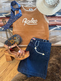 A styled flat-lay outfit featuring a tan knit sleeveless top with 'Rodeo' written in white script, paired with dark blue denim shorts, a blue patterned neck scarf, brown leather platform sandals, and a beige straw hat, arranged on a patterned rug over a wood floor.