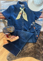 A styled outfit flat lay featuring a dark blue denim shirt and matching jeans, accessorized with a yellow patterned necktie, a blue embroidered crossbody bag, brown platform sandals, and a white straw hat, arranged on a patterned rug and a brown hide over a wood floor.