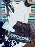 A styled flat-lay outfit featuring a cream short-sleeve t-shirt, a dark brown A-line skirt with a turquoise and black geometric pattern, a white low-top sneaker, a black ribbon, a green beaded necklace, and a baseball cap, arranged on a patterned rug and cowhide over a wood floor.