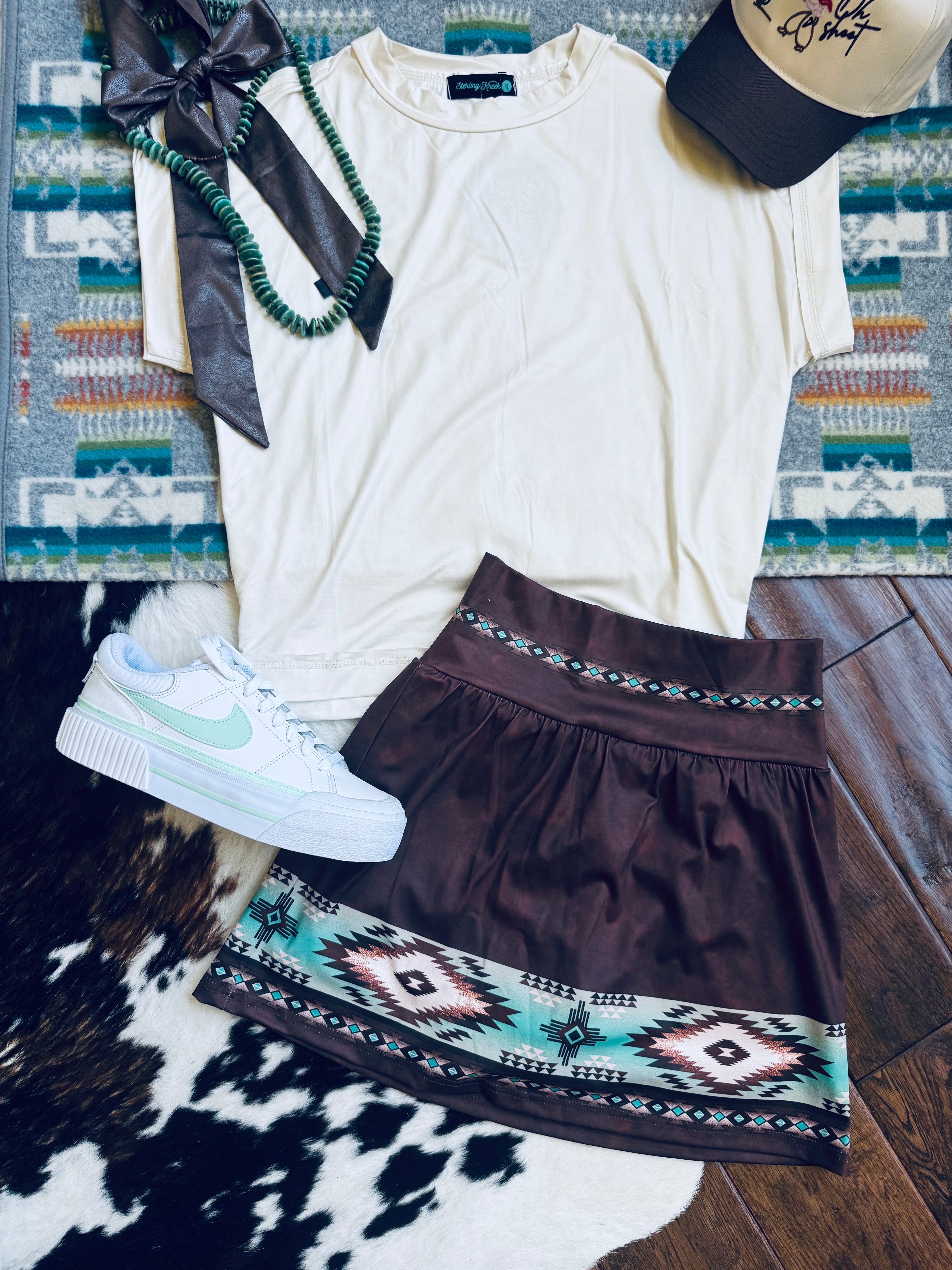A styled flat-lay outfit featuring a cream short-sleeve t-shirt, a dark brown A-line skirt with a turquoise and black geometric pattern, a white low-top sneaker, a black ribbon, a green beaded necklace, and a baseball cap, arranged on a patterned rug and cowhide over a wood floor.