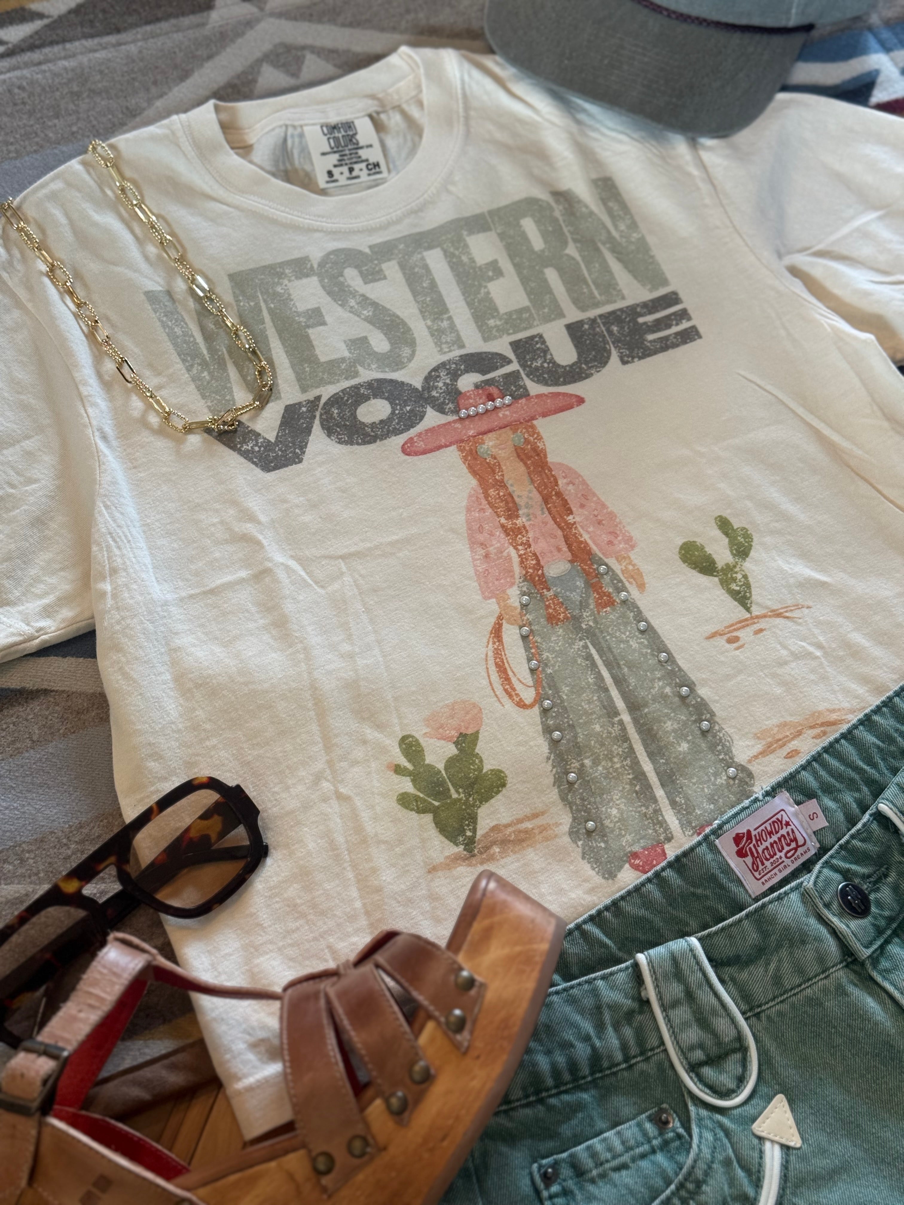 A cream-colored graphic t-shirt featuring a western-themed illustration and the text 'WESTERN VOGUE' in distressed gray letters. The shirt is styled in a flat lay with a gold chain necklace, sunglasses, brown leather sandals, and green denim shorts on a gray patterned fabric background.