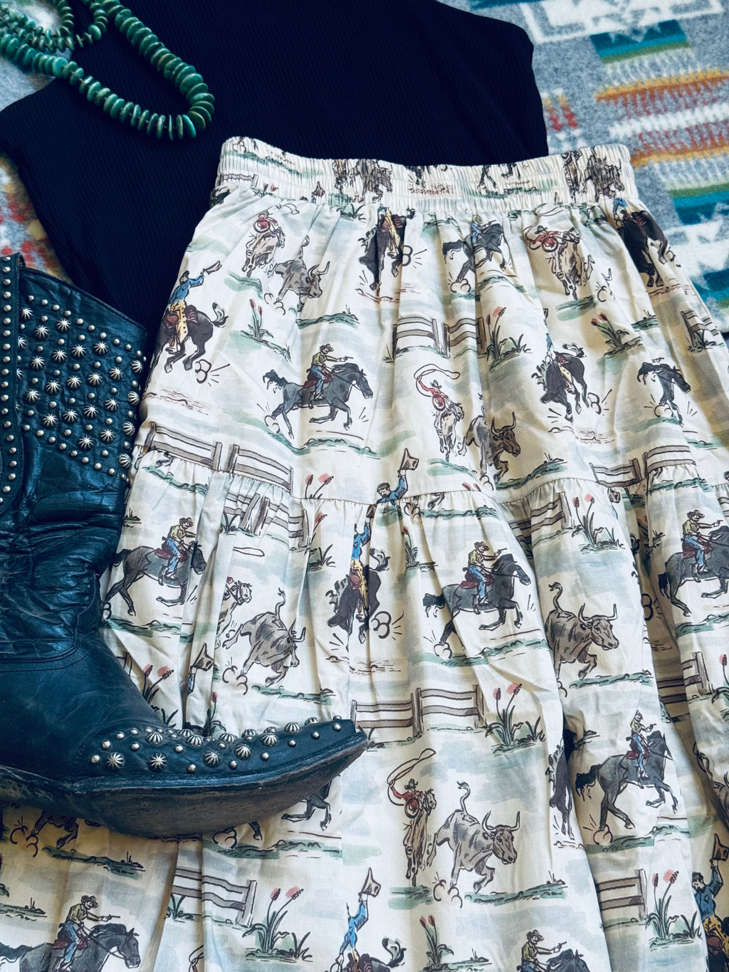 A close-up flat lay of a tiered skirt with an all-over western cowboy and cattle print on an off-white background, styled with a black studded boot, a black top, and a green beaded necklace.