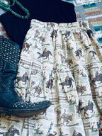 A close-up flat lay of a tiered skirt with an all-over western cowboy and cattle print on an off-white background, styled with a black studded boot, a black top, and a green beaded necklace.
