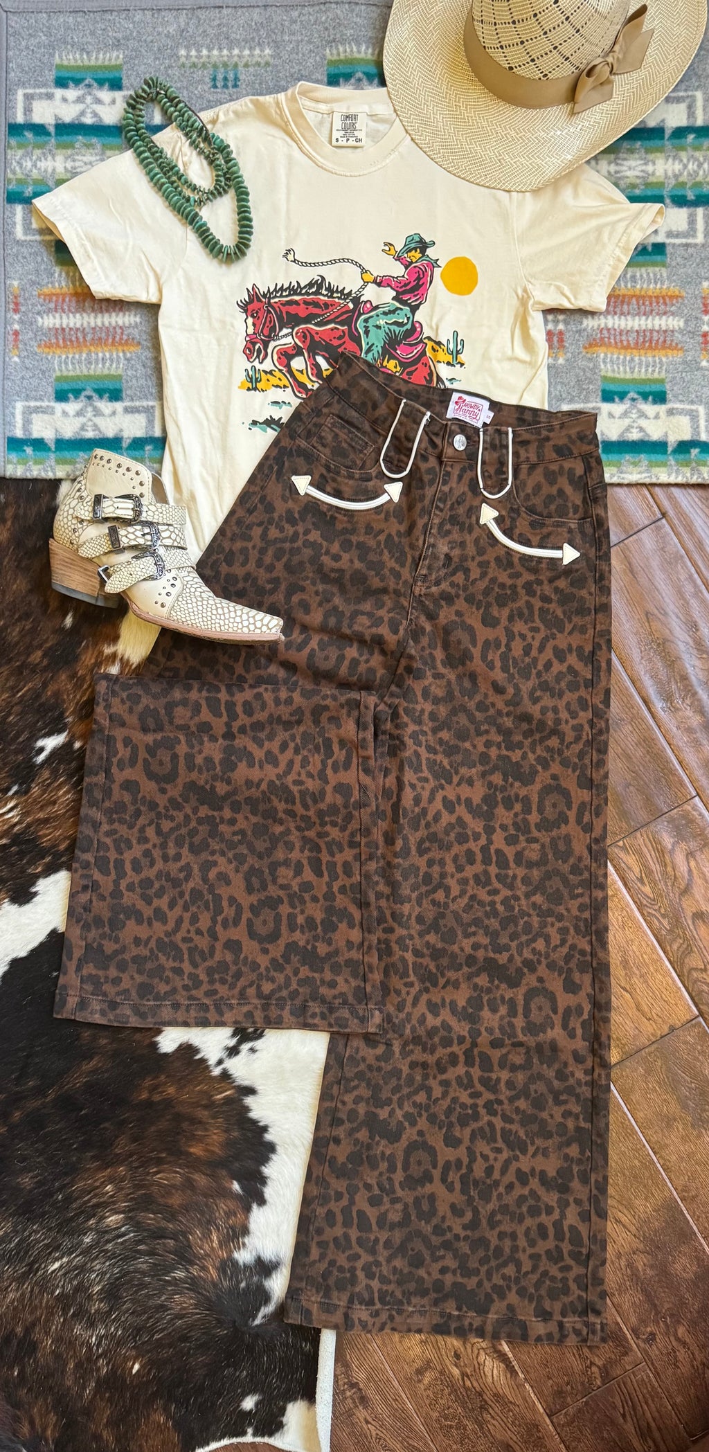 A styled flat-lay outfit featuring brown leopard print wide-leg pants, a cream graphic t-shirt, a straw cowboy hat, white western boots, a green beaded necklace, and a patterned rug on a wooden floor.