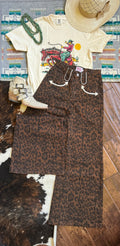 A styled flat-lay outfit featuring brown leopard print wide-leg pants, a cream graphic t-shirt, a straw cowboy hat, white western boots, a green beaded necklace, and a patterned rug on a wooden floor.