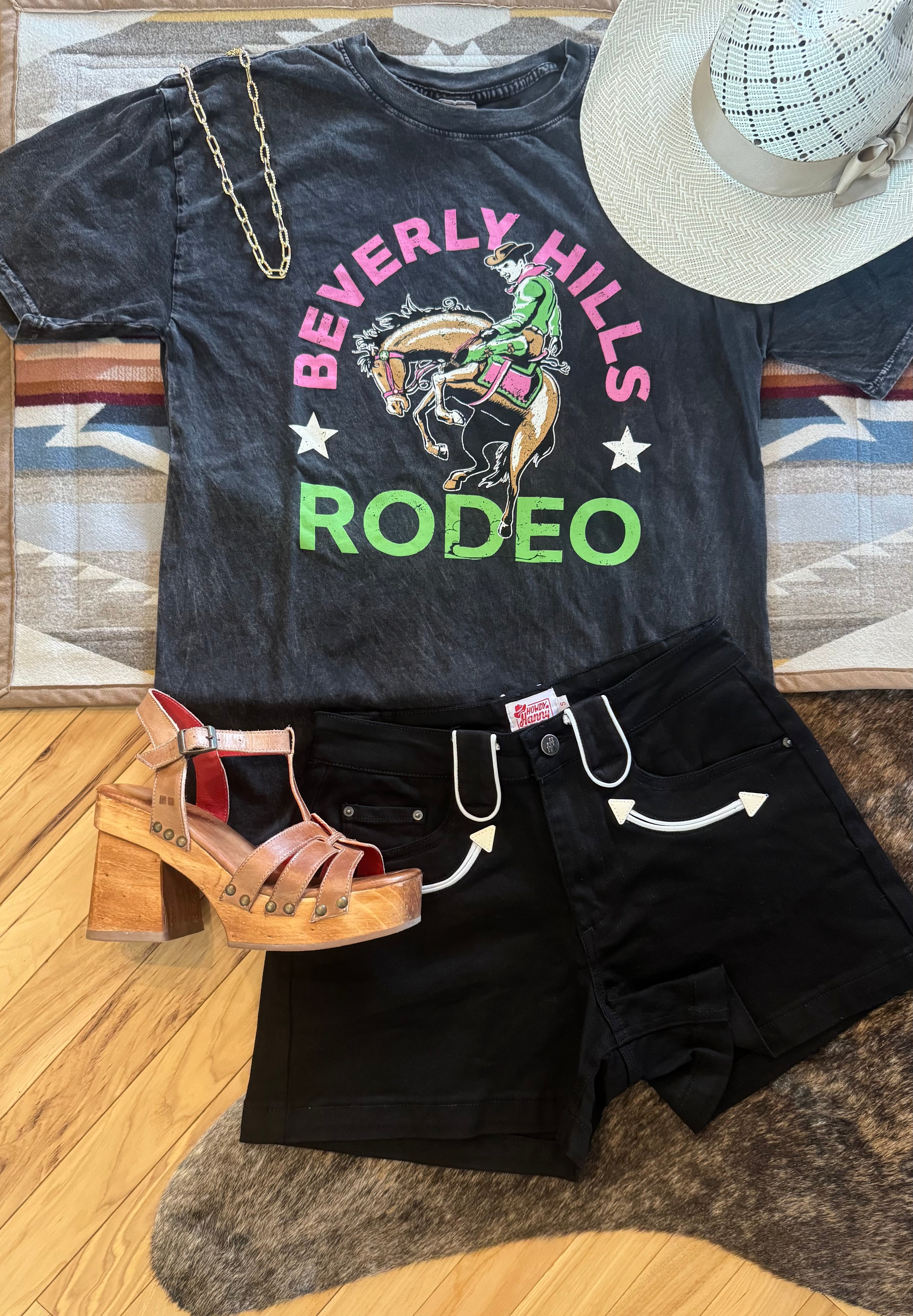 A styled outfit flat lay featuring a faded black graphic t-shirt with 'Beverly Hills Rodeo' text and a cowboy illustration, paired with black denim shorts, brown platform sandals, and a white straw hat on a patterned rug and wood floor.