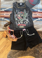 A styled flat-lay outfit featuring a dark gray graphic tank top with 'Wild Westeroo Ranch' text, black denim shorts, brown platform sandals, sunglasses, a gold chain necklace, and a gray baseball cap, arranged on a patterned rug over a wood floor.