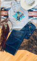 A styled flat-lay outfit featuring a light gray graphic tank top with a cowboy riding a bucking horse print, paired with dark blue denim jeans. The outfit is arranged on a striped southwestern-patterned rug alongside a brown leather fringe bag, a white straw hat, and a brown and black animal print rug over light wood flooring.