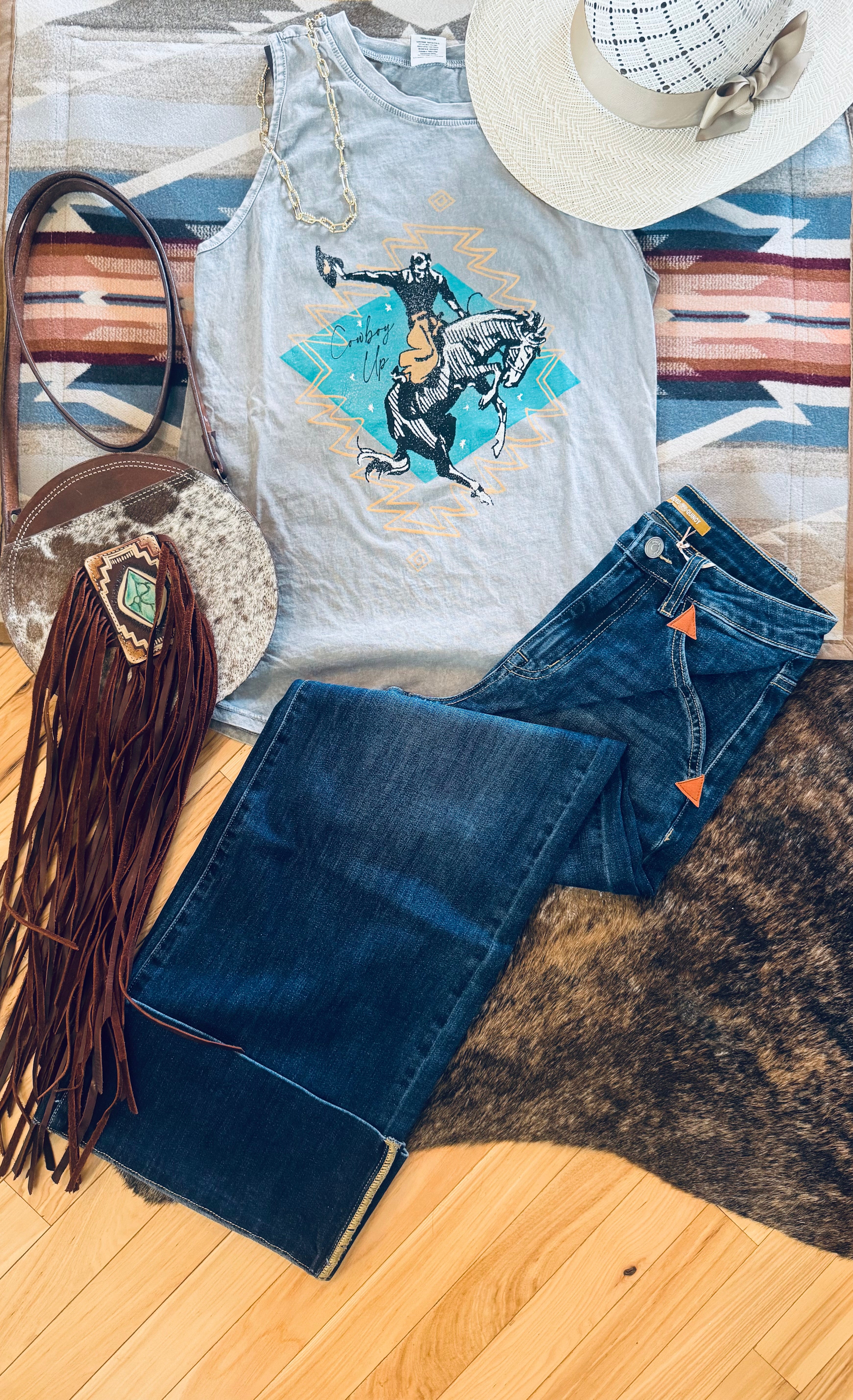 A styled flat-lay outfit featuring a light gray graphic tank top with a cowboy riding a bucking horse print, paired with dark blue denim jeans. The outfit is arranged on a striped southwestern-patterned rug alongside a brown leather fringe bag, a white straw hat, and a brown and black animal print rug over light wood flooring.