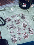 A sage green graphic t-shirt featuring a collage of vintage western and cowboy illustrations in brown and white. The shirt is styled flat-lay with a black coiled cord, a camouflage cap, and blue jeans.