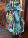 A teal necktie with a colorful floral print and a matching satin bow, displayed on a rustic cactus-shaped stand.