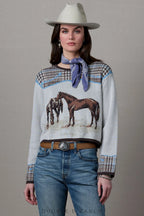 A model wearing a light gray knit sweater with a horse graphic, patterned houndstooth shoulder and cuff details, a blue neck scarf, blue jeans, and a white cowboy hat, posed against a neutral gray background.