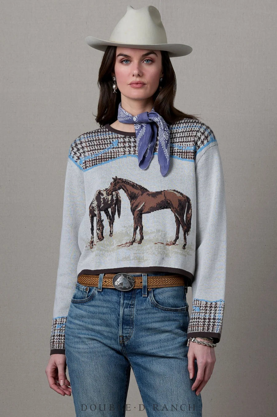 A model wearing a light gray knit sweater with a horse graphic, patterned houndstooth shoulder and cuff details, a blue neck scarf, blue jeans, and a white cowboy hat, posed against a neutral gray background.