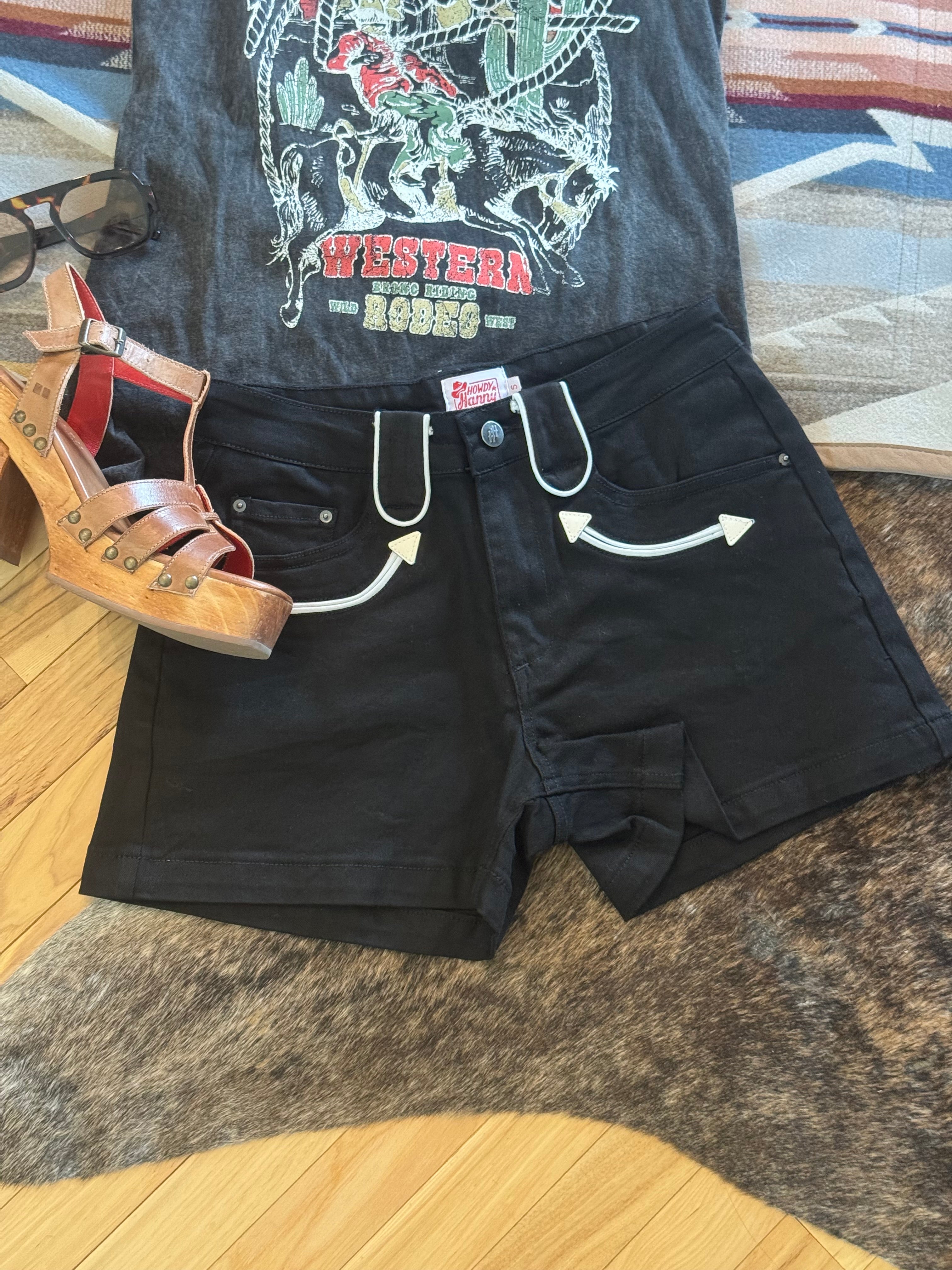 Black denim shorts with white graphic arrow details on the pockets, styled in a flat lay with a graphic t-shirt, brown platform sandals, and glasses on a patterned rug and wood floor.