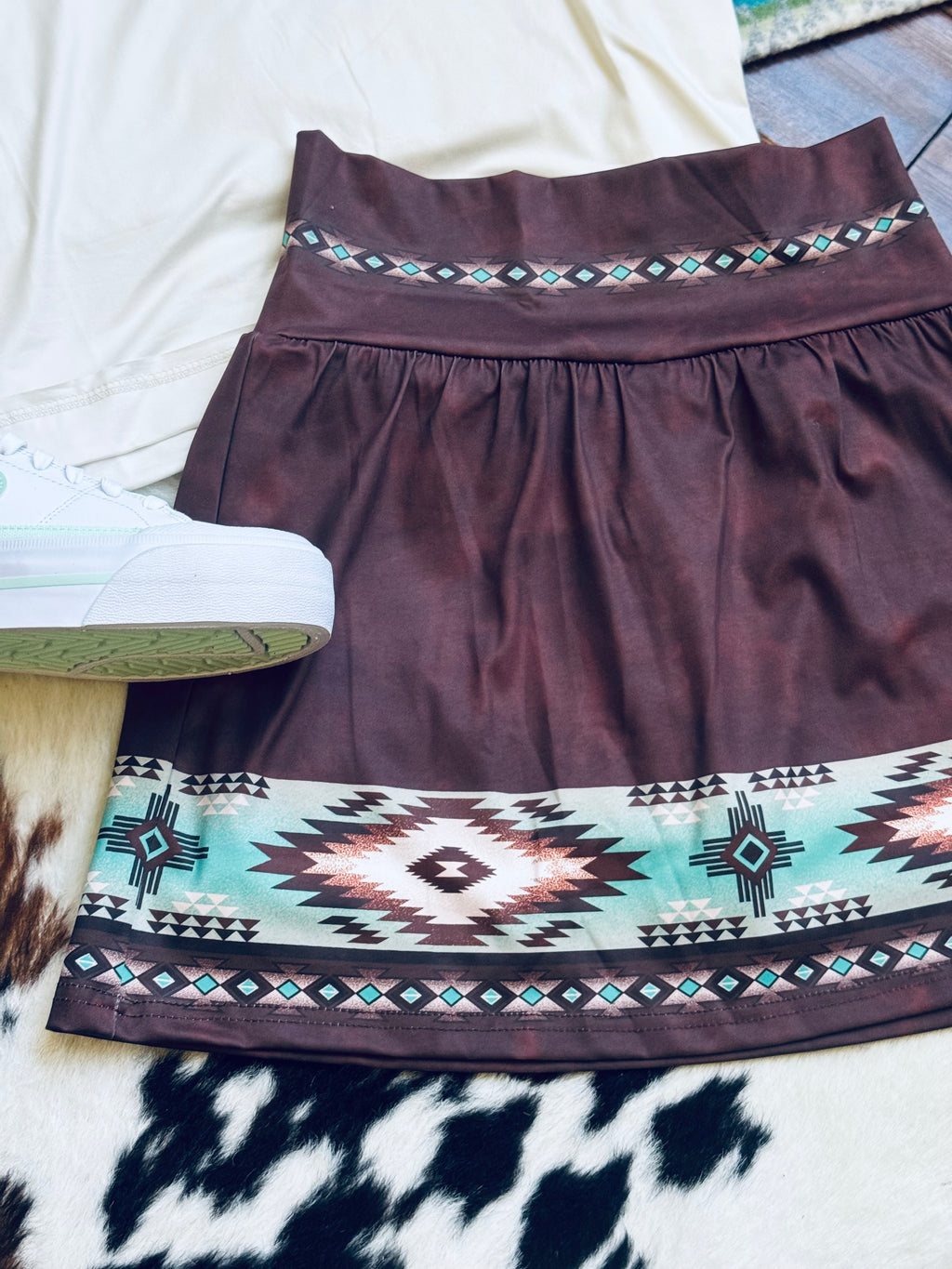 A dark brown A-line skirt/skort with a wide Southwestern geometric pattern border, styled flat-lay on a white surface with a white sneaker partially visible.