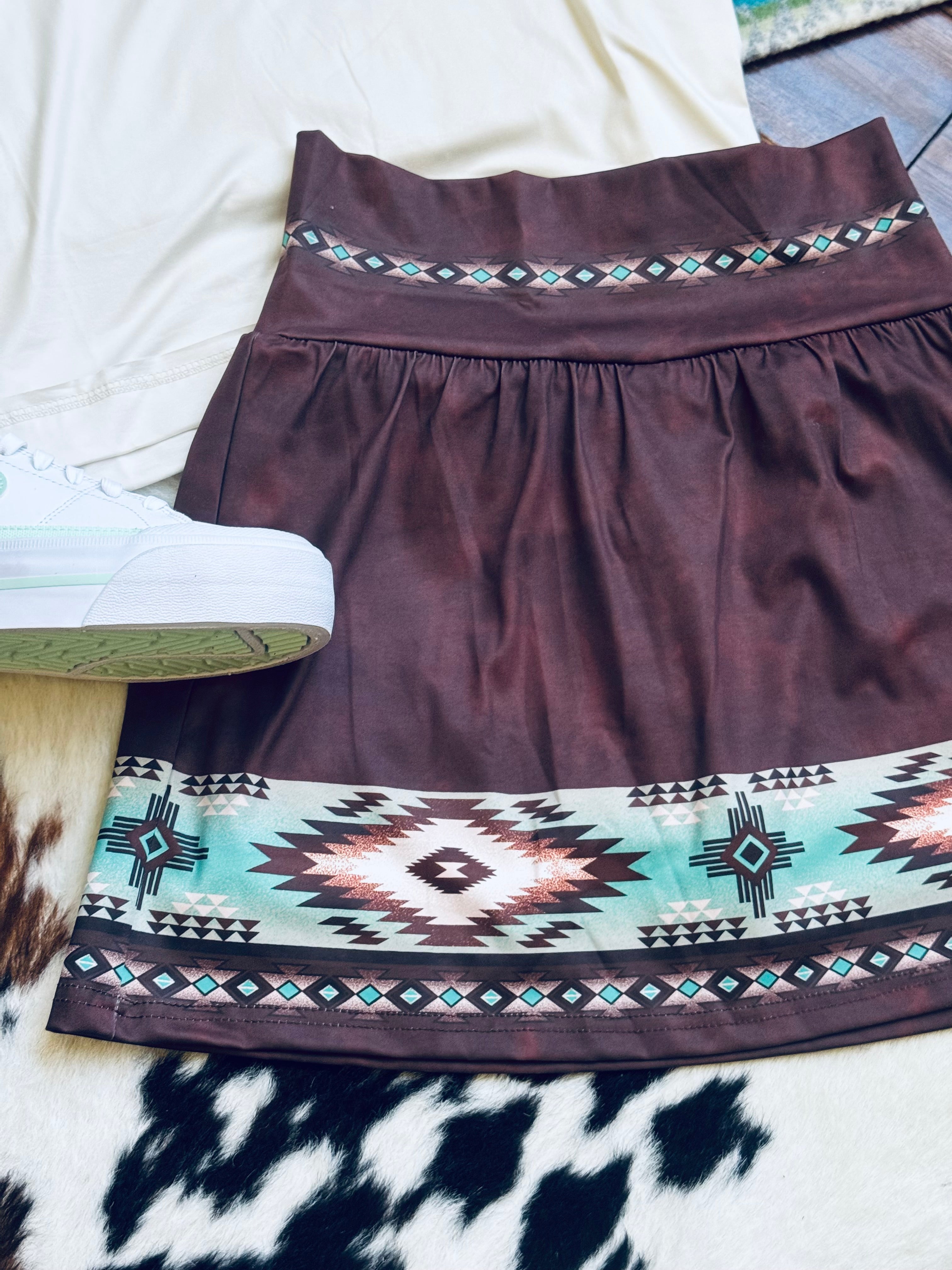 A dark brown A-line skirt/skort with a wide Southwestern geometric pattern border, styled flat-lay on a white surface with a white sneaker partially visible.