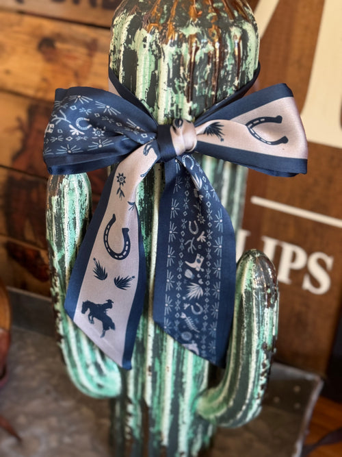 A navy blue and cream bandana bow with western-themed prints, tied around a distressed green cactus display in a rustic indoor setting.