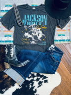 A styled flat-lay featuring a dark charcoal graphic t-shirt with 'Jackson Hole Rodeo' and a cowboy riding design, paired with blue jeans, a black cowboy boot, a black hat, and a beaded necklace on a patterned rug over a wood floor.