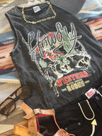 A black washed sleeveless graphic top with a western design featuring a cowboy, cactus, and the text 'Western Texas 1969', styled flat-lay with black shorts, sunglasses, a gold chain, and a belt on a patterned rug.