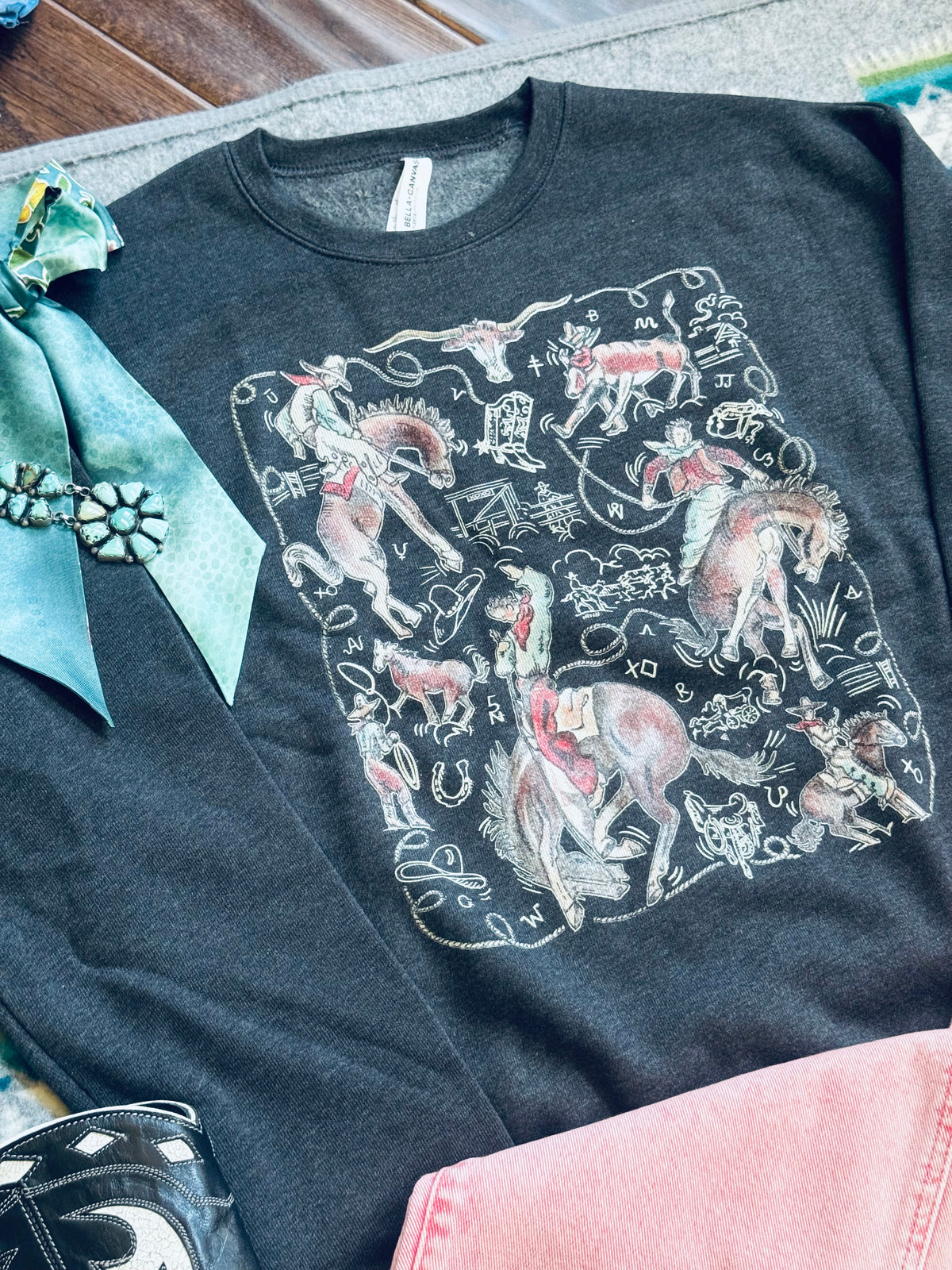 A dark charcoal gray graphic t-shirt featuring a western-themed print of cowboy riders on horseback, lasso lines, and zodiac symbols. The shirt is styled in a flat lay with a teal bow, a black western boot, and pink denim on a gray fabric background.