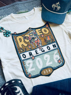 A white graphic T-shirt laid flat with a large western-style 'Rodeo Oregon 2026 USA' print on the front. The shirt is styled with a blue western cap, dark denim shorts, and turquoise jewelry on a gray fabric background.
