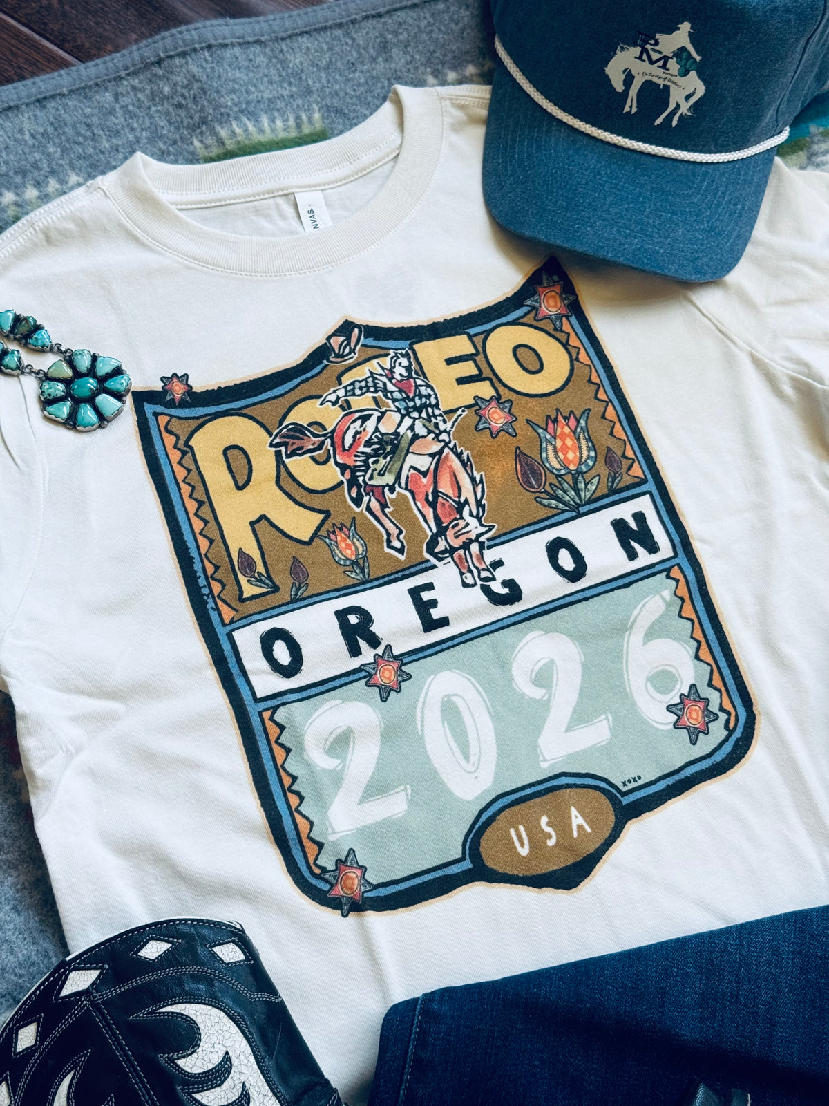 A white graphic T-shirt laid flat with a large western-style 'Rodeo Oregon 2026 USA' print on the front. The shirt is styled with a blue western cap, dark denim shorts, and turquoise jewelry on a gray fabric background.