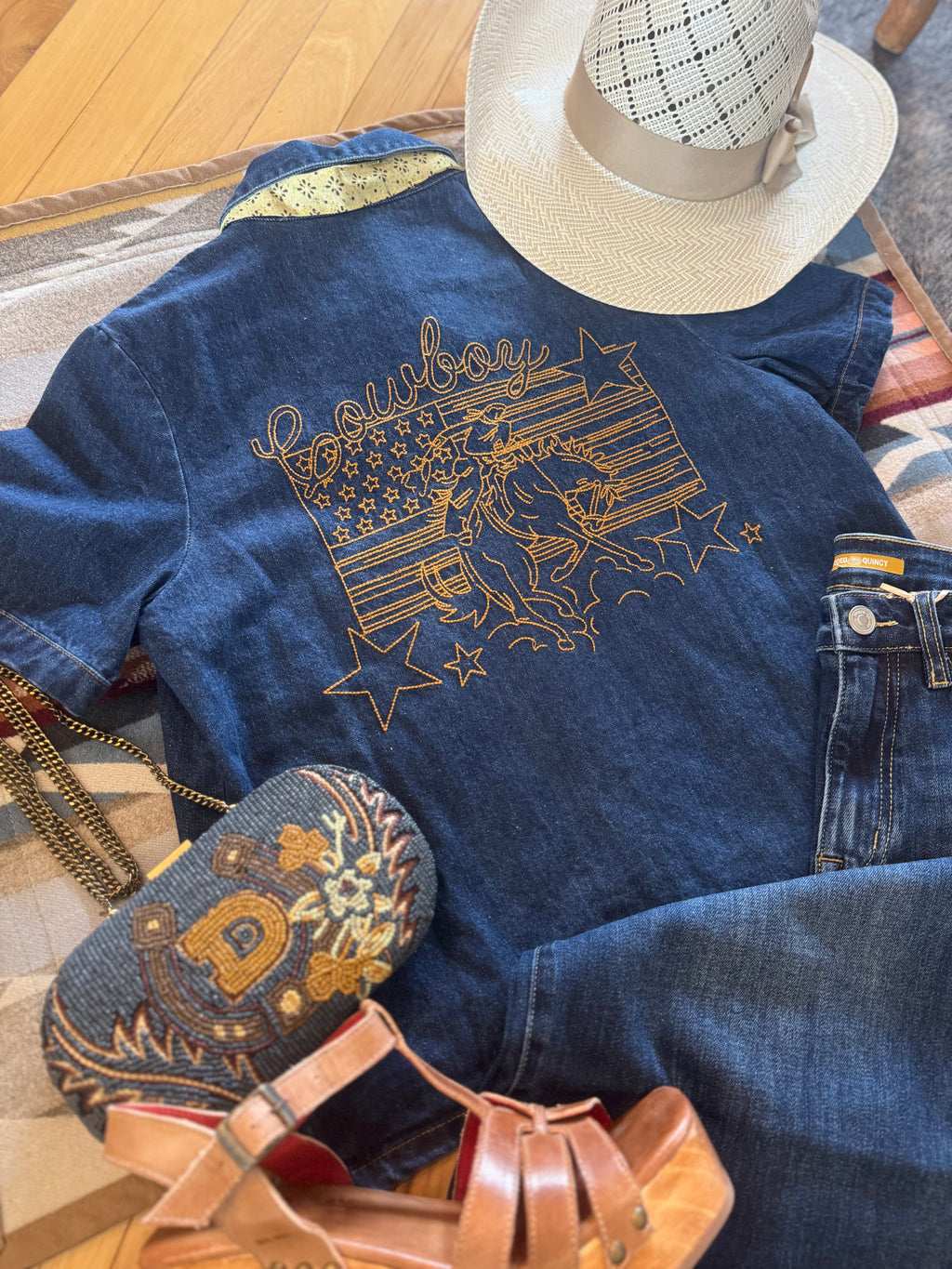 A styled flat-lay of a blue denim shirt with gold western embroidery reading 'Cowboy' and an American flag motif, paired with blue jeans, a white cowboy hat, a patterned clutch, and tan platform sandals on a patterned rug over a wood floor.
