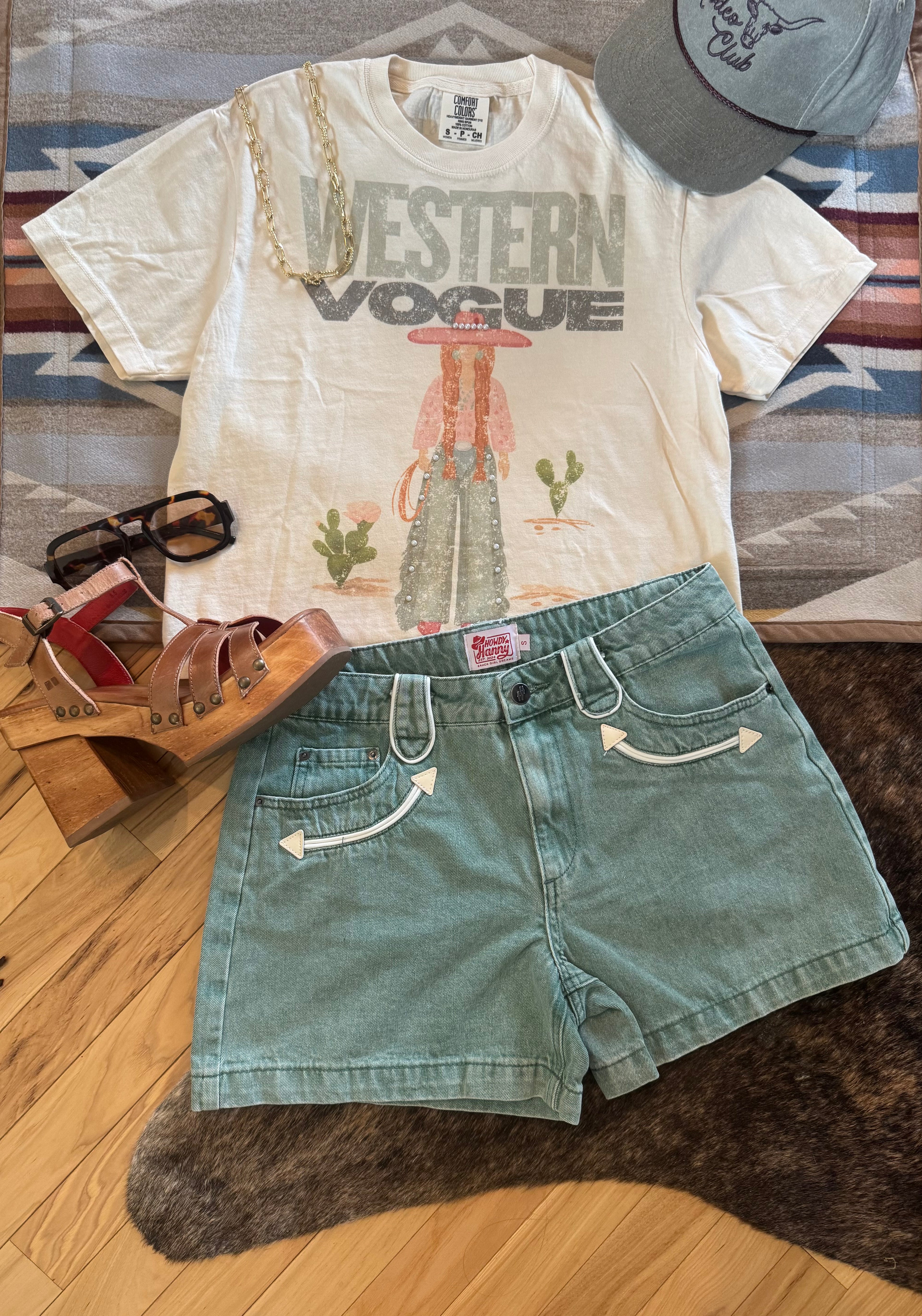 A styled flat-lay outfit featuring a cream graphic t-shirt with 'WESTERN VOGUE' text and a western print, paired with sage green denim shorts with embroidered arrow details. The outfit is styled with a gray baseball cap, brown platform sandals, and black sunglasses on a patterned rug and wood floor.