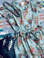 A light sage green western-style shirt dress with a colorful cowboy and horse print, styled flat-lay with a black coiled necklace and a black western boot on a patterned rug.