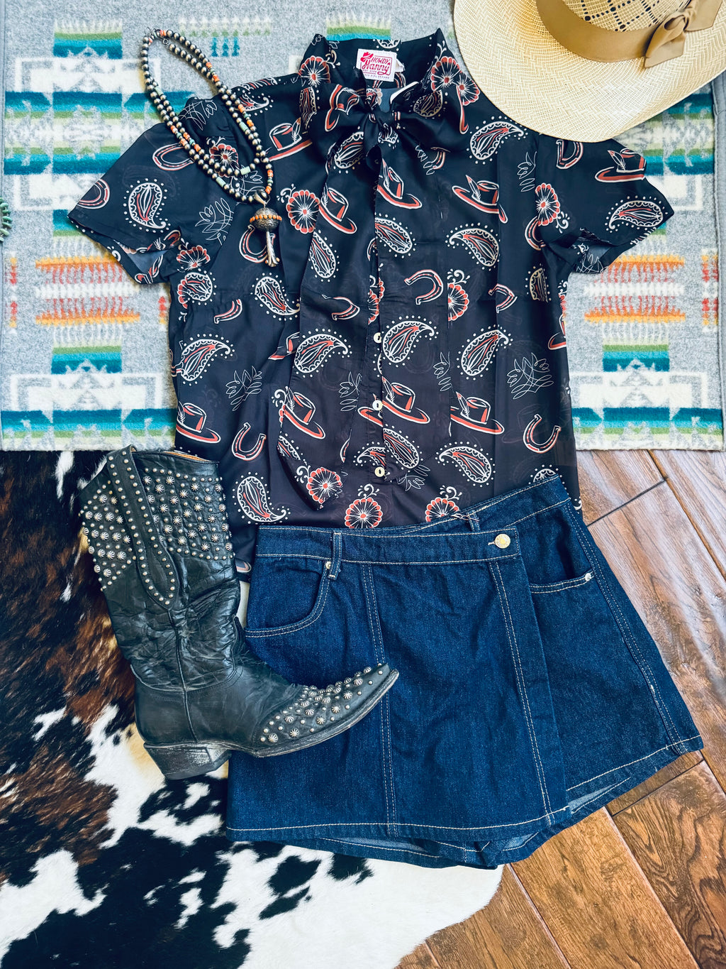 A styled flat-lay of a women's western outfit featuring a black short-sleeve button-up shirt with a paisley and horseshoe print, a dark blue denim mini skirt, a black studded cowboy boot, a beaded necklace, and a straw hat, arranged on a patterned rug and wood floor.