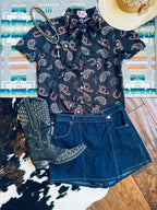 A styled flat-lay of a women's western outfit featuring a black short-sleeve button-up shirt with a paisley and horseshoe print, a dark blue denim mini skirt, a black studded cowboy boot, a beaded necklace, and a straw hat, arranged on a patterned rug and wood floor.