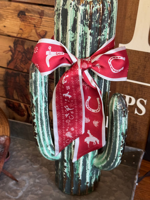 A distressed green cactus-shaped decorative object tied with a red and white western-themed ribbon featuring white horseshoe and rider motifs, set against a rustic wood background.