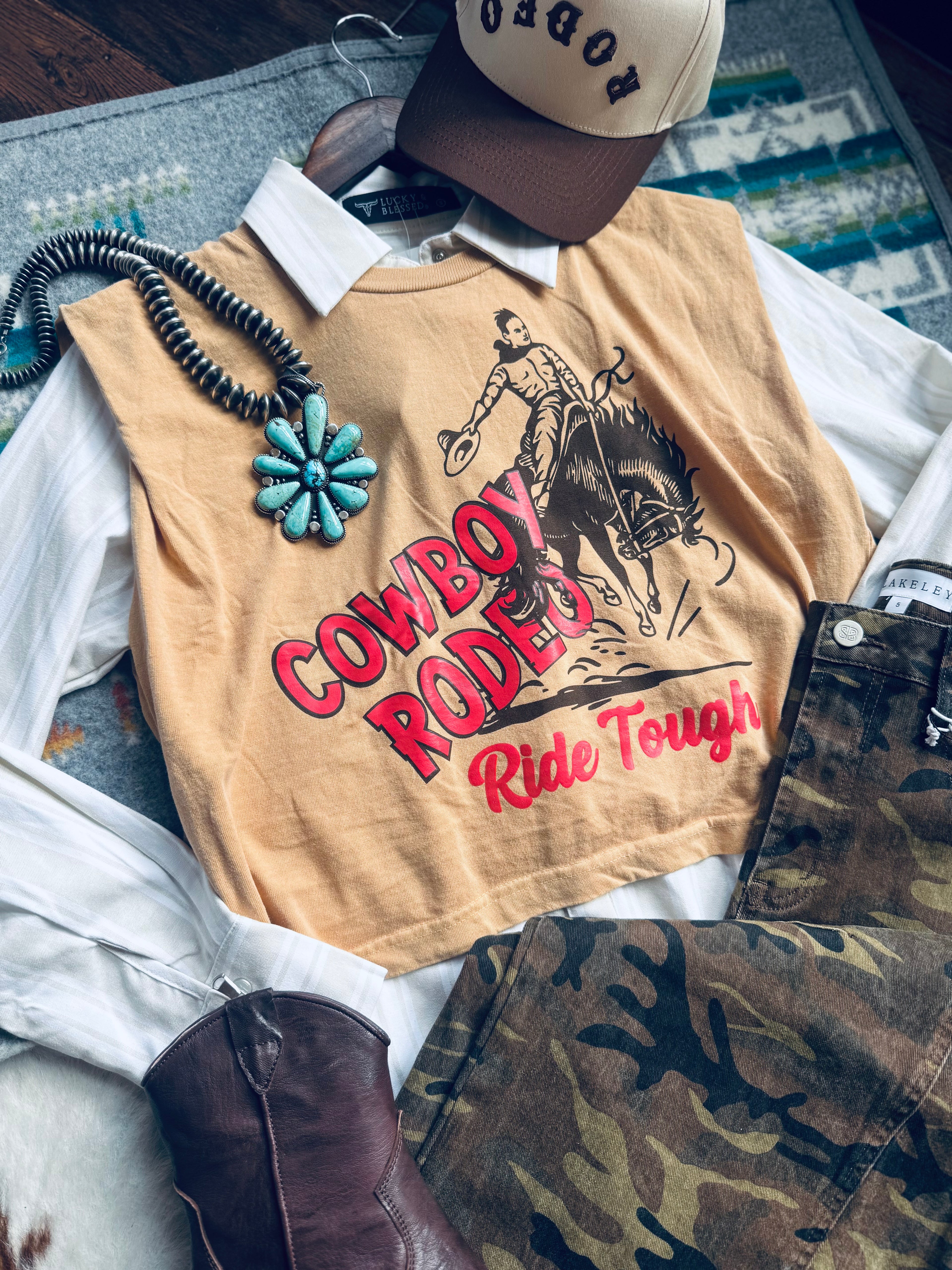 Beige shirt with cowboy graphic and text, surrounded by camouflage pants and a cap on a textured surface.