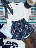 A styled flat-lay outfit featuring a cream short-sleeve t-shirt, navy blue patterned shorts with a geometric diamond design, a white Nike sneaker, a black ribbon, a green beaded necklace, and a baseball cap, arranged on a patterned rug and wood floor.