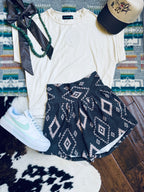 A styled flat-lay outfit featuring a cream short-sleeve t-shirt, navy blue patterned shorts with a geometric diamond design, a white Nike sneaker, a black ribbon, a green beaded necklace, and a baseball cap, arranged on a patterned rug and wood floor.