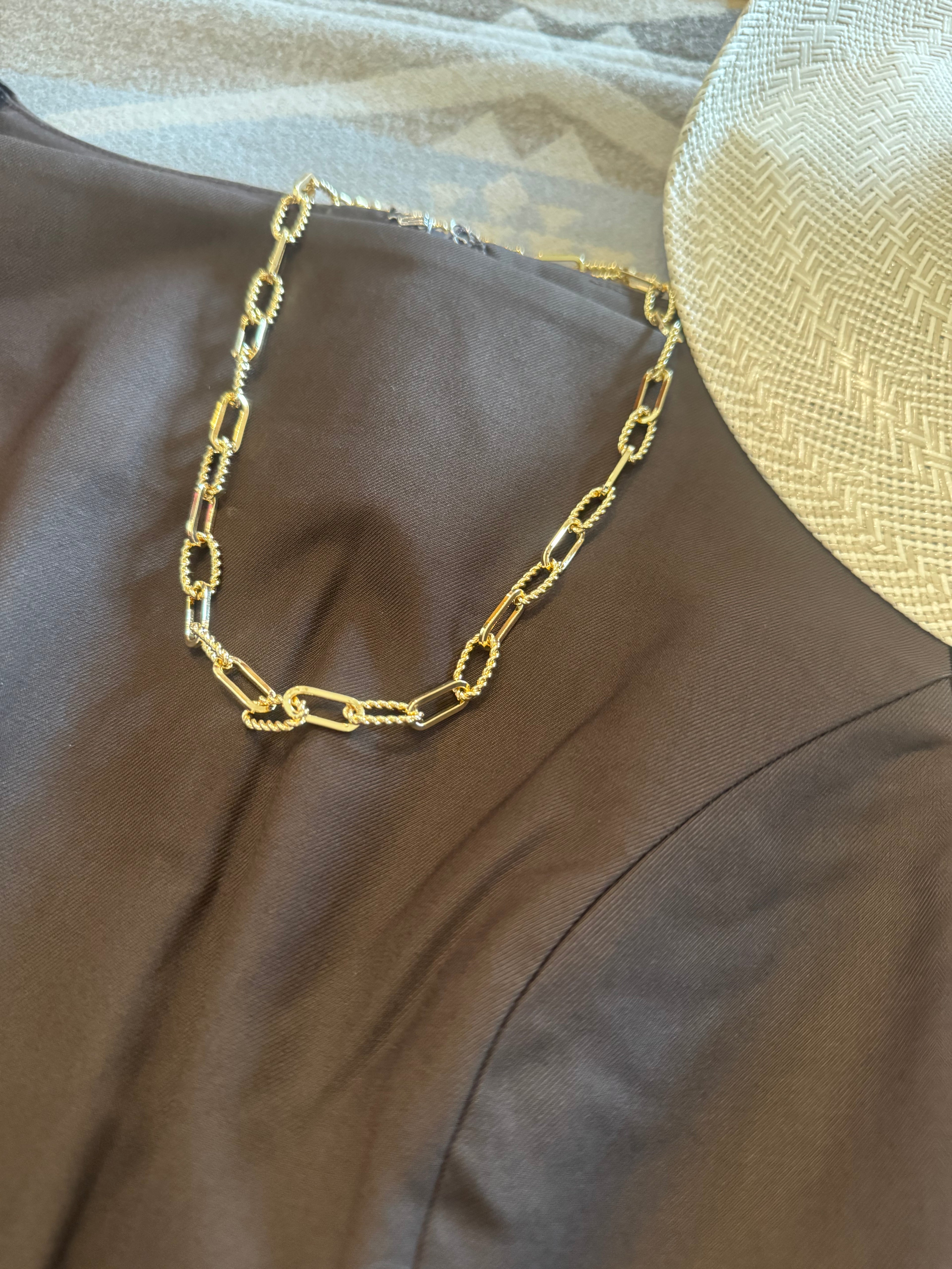 A gold paperclip chain necklace draped over a brown satin fabric, with a woven straw hat partially visible in the upper right corner.