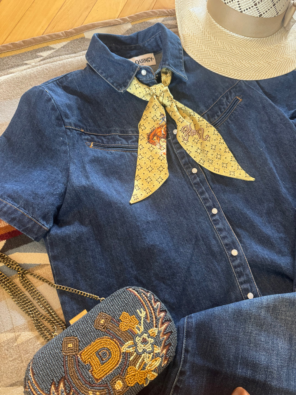 A styled flat-lay of a blue denim button-up shirt with a yellow patterned neck scarf, a matching embroidered denim crossbody bag, and a straw hat on a patterned rug.