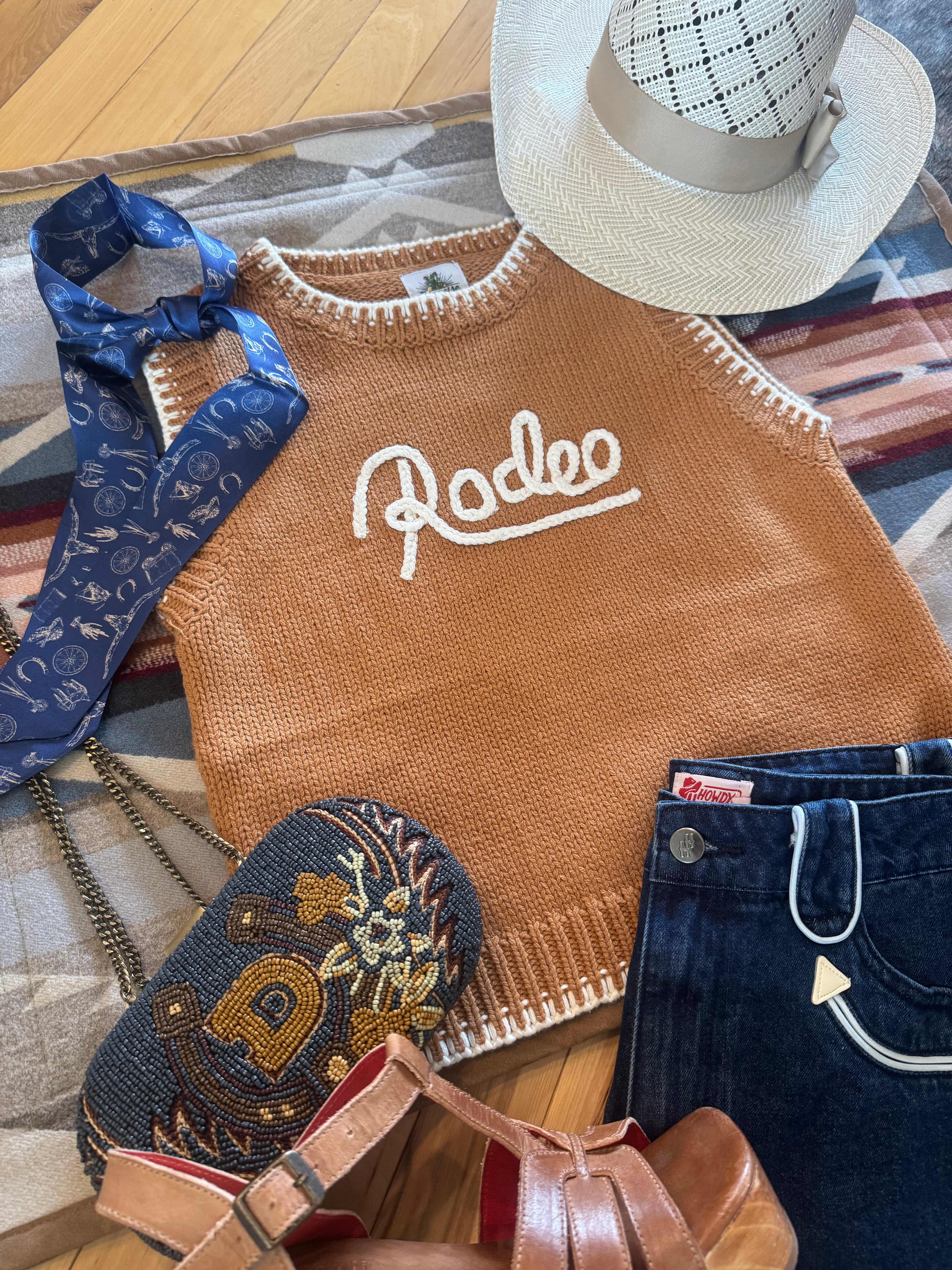 A styled flat-lay featuring a brown knit sweater vest with white 'Rodeo' embroidery, styled with a white cowboy hat, blue jeans, a blue patterned silk tie, a blue embroidered pouch, and brown leather sandals on a patterned rug over a wooden floor.