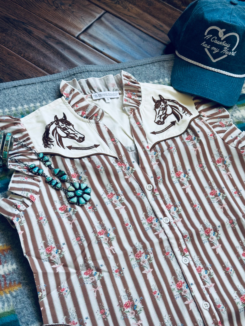 A western-style striped button-up shirt laid flat on a patterned rug with a wooden floor in the background. The shirt has vertical brown and cream stripes, a small floral print, and two cream pointed collar panels with embroidered horse heads.