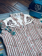 A western-style striped button-up shirt laid flat on a patterned rug with a wooden floor in the background. The shirt has vertical brown and cream stripes, a small floral print, and two cream pointed collar panels with embroidered horse heads.